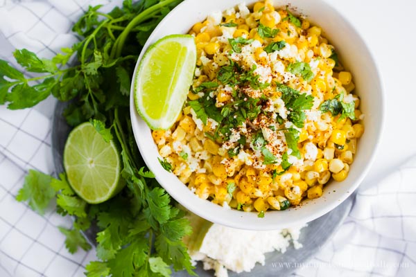 A white bowl full of grilled corn topped with cheese, chopped cilantro and lime wedges on the side.