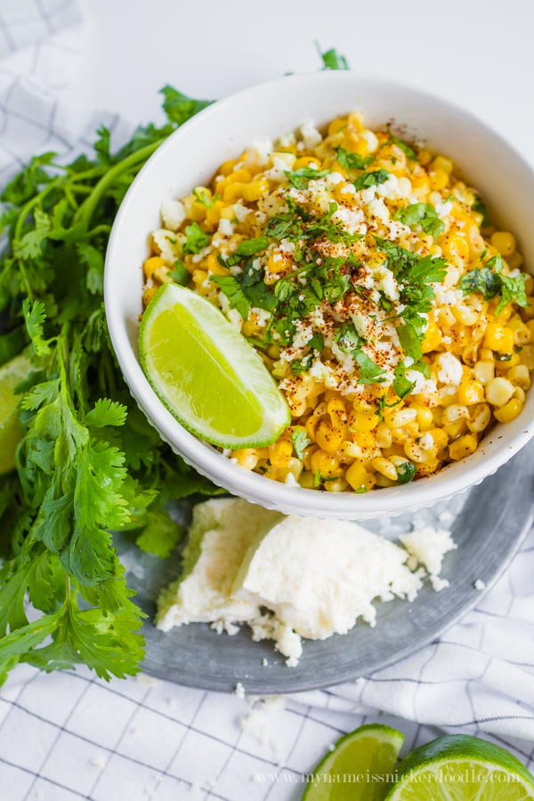 A white bowl full of grilled corn topped with cheese, chopped cilantro and lime wedges on the side.