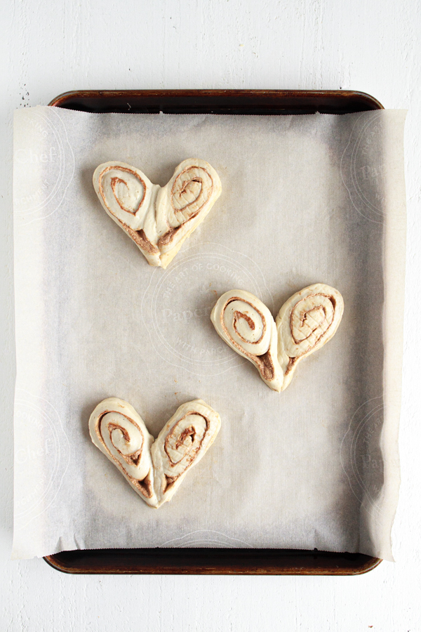 Three heart shaped cinnamon rolls on a parchment lined baking sheet.