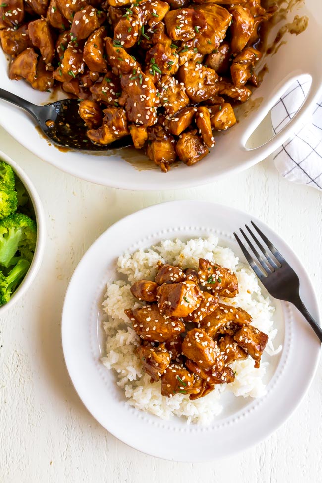 A white bowl with sesame chicken and a plate of rice.