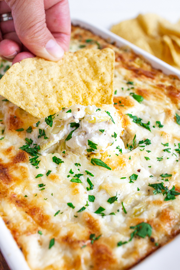 Tortilla chip being dipped into hot and cheesy artichoke dip.